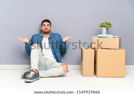 Handsome young man moving in new home among boxes having doubts with confuse face expression