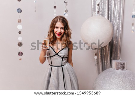  Girl 25 years in beautiful bright dress with glass of champagne in hands, having fun in front of camera on eve of Christmas holidays