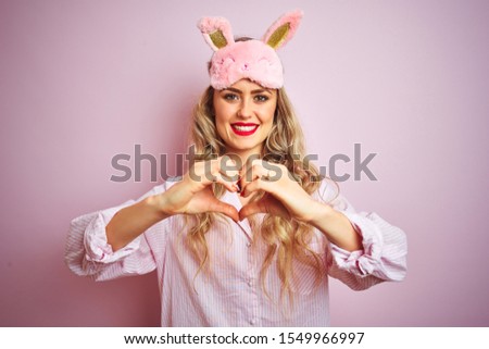 Young beautiful woman wearing pajama and sleep mask over pink isolated background smiling in love doing heart symbol shape with hands. Romantic concept.