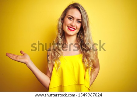 Young attactive woman wearing t-shirt standing over yellow isolated background smiling cheerful presenting and pointing with palm of hand looking at the camera.