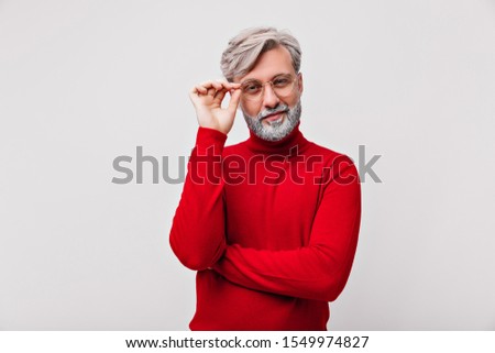 Joyful old man looking through the glasses with interest. Studio photo of pleased male model in red sweater.
