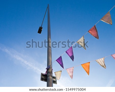 Multi-coloured bunting tied to a lamp post, shot in colour from below against a blue sky with light wispy clouds. 