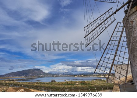 Sicilian landscape with mountain and city Erice in background and blades or sails of old stone windmill in traditional salt production in Saline di Trapani. Picture taken in summer cloudy sunny day.