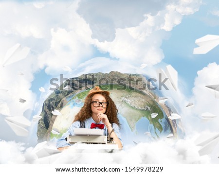 Side view of hard-working female writer using typing machine while sitting at the table with flying paper planes and Earth globe among cloudy skyscape on background. Elements of this image furnished