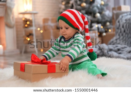 Little baby with elf hat and Christmas gift on floor at home