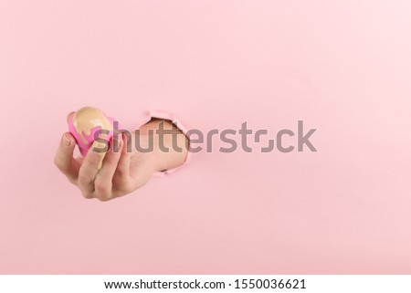 Girl hand holds beauty blender in foundation, concealer from a hole in a pink background. Makeup artist concept, copy space.
