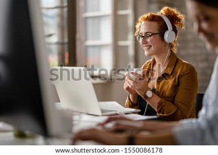 Happy female entrepreneur wearing headphones while surfing the net on laptop and drinking coffee in the office. 