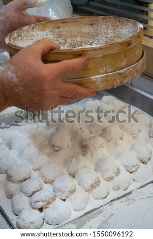 Pastry chef's hands sifting powder sugar with a wooden sieve over a plate of kourabiedes during preparation. Scene from working in bakery/pastry shop
