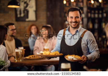 Happy waiter holding plates with food and looking at camera while serving guests in a restaurant.  Royalty-Free Stock Photo #1550134583