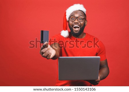 Portrait of an excited young african american man dressed in santa claus costume standing isolated over red background, holding laptop computer, showing plastic credit card.