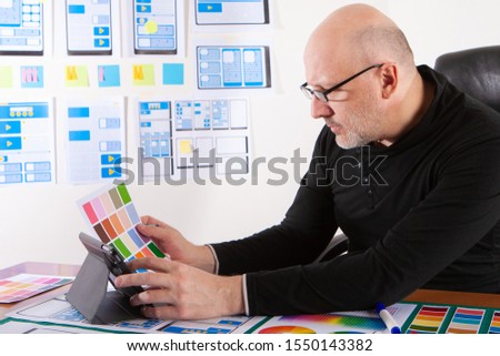 The work of the designer of mobile applications. The man in glasses works with a color palette and a tablet. Select a color scheme for the user interface. Program display color is selected.