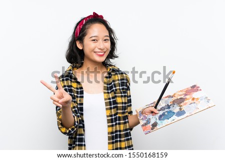 Young artist woman holding a palette over isolated white background smiling and showing victory sign