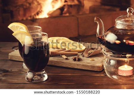 Two glasses of mulled wine and teapot on a wooden table in front of a burning fireplace.