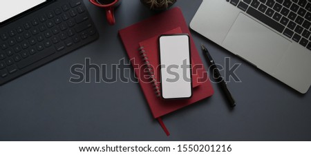 Overhead shot of dark modern workplace with blank screen smartphone and office supplies on grey desk background