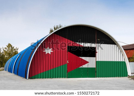 Close-up of the national flag of Jordan
 painted on the metal wall of a large warehouse the closed territory against blue sky. The concept of storage of goods, entry to a closed area, logistics
