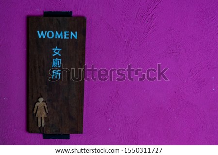 Toilet sign with icon of woman and the word "women" in English and Chinese language on wooden panel against vibrant pink purple background.