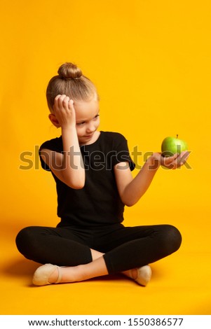 Full body little girl in leotard scratching head and looking at green apple while sitting against yellow background during break in training