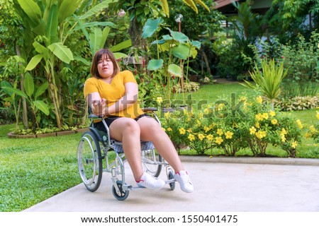 Painful overweight young woman sitting alone on the wheelchair while her hands are sinking due to nervous system illness, hemiplegia and paralysis in the garden. Healthy concept. Royalty-Free Stock Photo #1550401475
