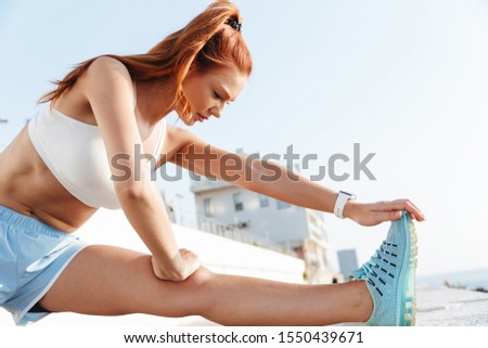 Image of attractive young woman in sportswear stretching her body while doing workout by seaside in morning