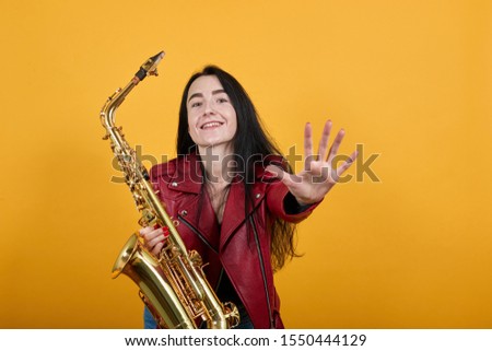 Portrait of crazy young woman in red casual clothes looking camera, keeping saxophone and smiling, standing with outstretched hands isolated on orange background. People lifestyle concept