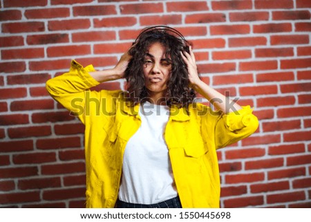 Positive human emotions. Portrait of happy emotional teenage girl with headphones, closed eyes, kiss to the camera. Wearing bright yellow cotton jacket and white shirt. Brick wall background.