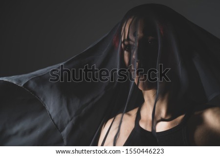 Woman in a black veil with dark makeup and an earring in her nose. Halloween Dead Bride Costume. Portrait of an ominous witch in front of a coven in a dark studio.