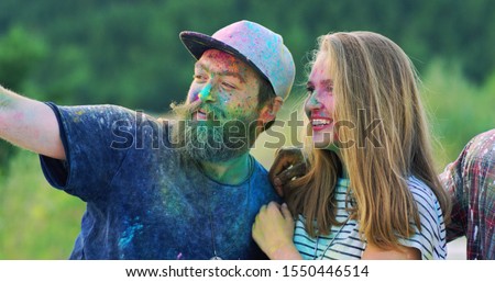 Caucasian cheerful young guy and girl in colorful paints smiling and posing to the camera of smartphone outdoor during holi fest. Taking selfie photos.