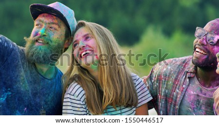 Close up of the joyful young mixed-races male and female friends laughing and celebrating holi holiday outdoors while posing to the smartphone camera and taking selfies photos.