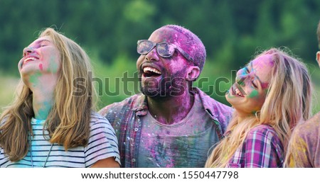 Friendly young multiethnic happy guys and girls in colorful paints smiling and posing to the camera of smartphone outside during holi fest. Taking selfie photos.