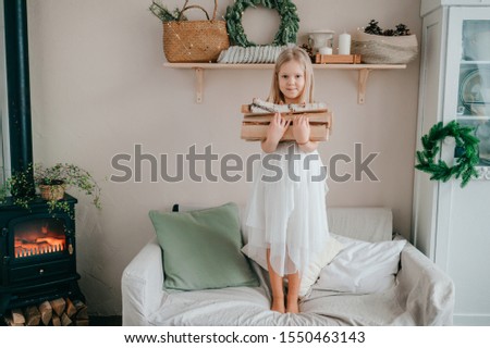 Beautiful little girl in white dress standing on bed with woods in her hands in chalet style room with fireplace