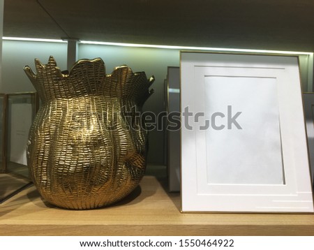 Gold metal vase with a textured antique finish and Thin gold frame with double passe-partout on a wooden shelf in a store. Ideas for a modern interior. Fashion decor