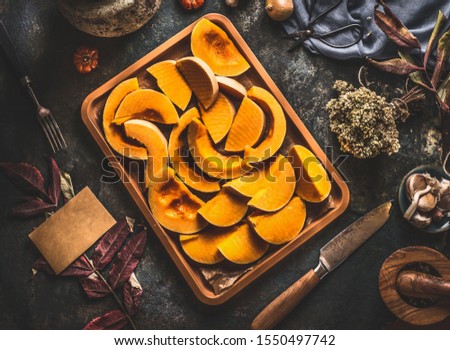 Pumpkin slices on baking sheet on dark rustic kitchen table background with knife and ingredients, top view. Pumpkin cooking recipes