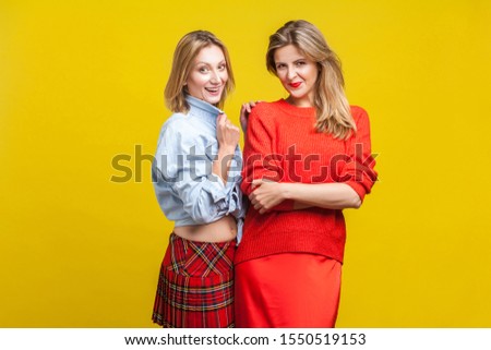 Best friends. Portrait of two fashion female models in stylish casual clothes standing together, smiling at camera. women showing autumn trends. indoor studio shot isolated on yellow background