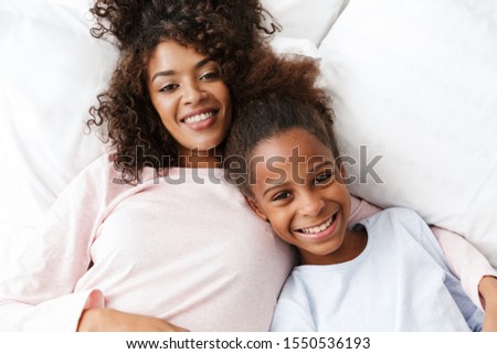 Image of cheerful african american woman and her daughter smiling and lying in bed at home