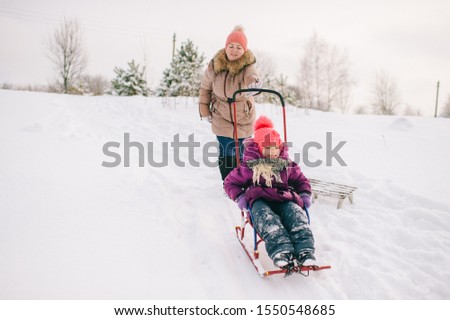 Mother having fun with her daughter sitting on sled in snowy winter day outdoors.