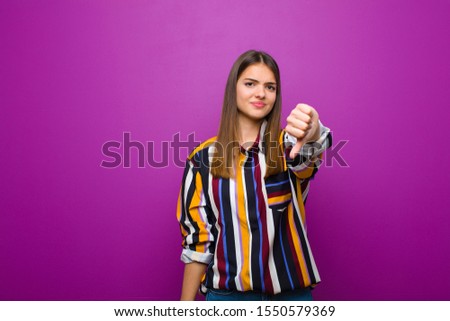young pretty woman feeling cross, angry, annoyed, disappointed or displeased, showing thumbs down with a serious look against purple background