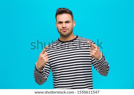 young hispanic man with a bad attitude looking proud and aggressive, pointing upwards or making fun sign with hands against blue background