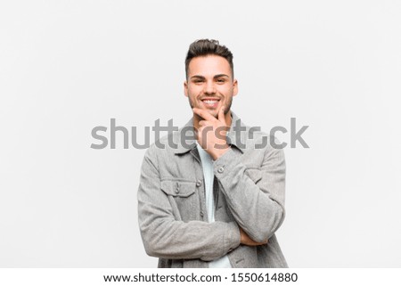 young hispanic man smiling, enjoying life, feeling happy, friendly, satisfied and carefree with hand on chin against gray background