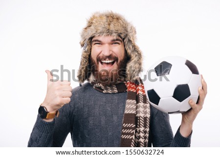 Cheerful bearded hipster man holding soccer ball and showing thumbs up