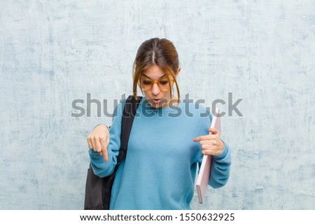 young student woman feeling shocked, open-mouthed and amazed, looking and pointing downwards in disbelief and surprise against grunge wall background
