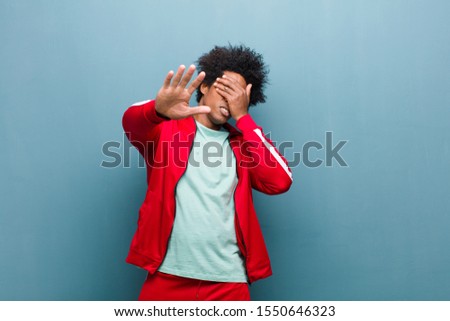 young black sports man covering face with hand and putting other hand up front to stop camera, refusing photos or pictures against grunge wall