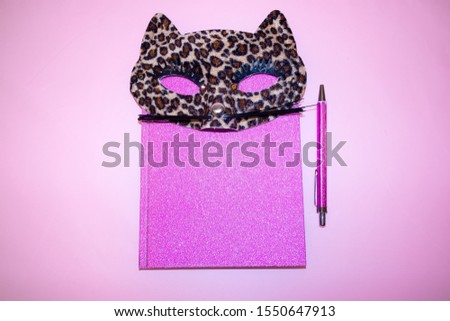 A pink diary and notebook with a cute masquerade cat mask in leopard pattern. Pink background. Flat lay view with copy space.