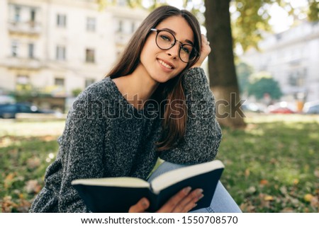 Image of beautiful smiling young woman wearing sweater and transparent eyeglasses, sitting outdoor iin the city park, reading the book. Young female student learning in the city street. 