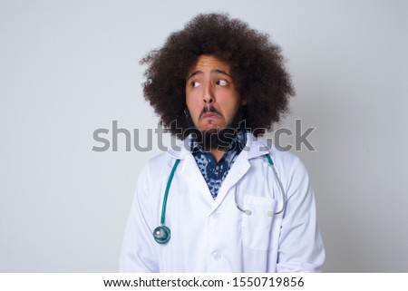 doctor man with snobbish expression curving lips and raising eyebrows, looking with doubtful and skeptical expression, suspect and doubt. Standing indoors over gray background.