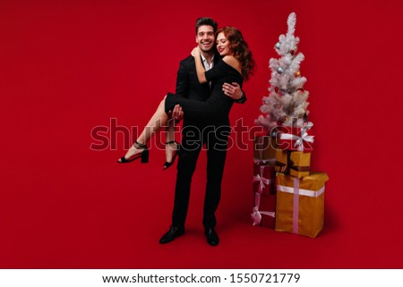 Photo of couple of lovers on red background decorated for new year. Man in suit holds his lady in his arms