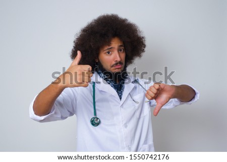 Pretty young pensive doctor man making good-bad sign. Displeased and unimpressed wearing casual clothes and standing against gray wall.