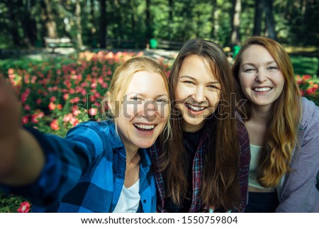 Happy young girls have fun laughing and taking selfies on a smartphone in the park on the background of green foliage and flowers. Blonde, brunette and girl with red hair, female friendship concept.