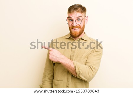 young red head man looking excited and surprised pointing to the side and upwards to copy space against beige background