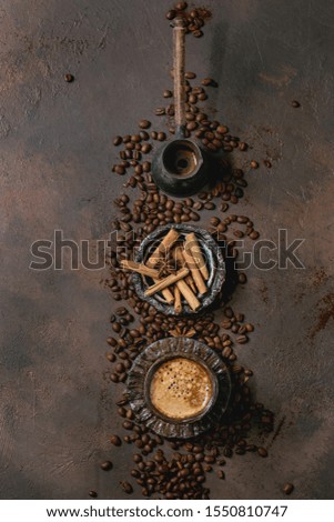 Black coffee espresso with foam in black ceramic cup, with saucer, cezve coffee pot, spices and roasted beans above over brown texture background. Flat lay, space