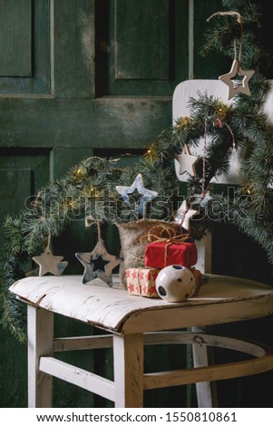 Christmas decoration fir tree branches, ceramic stars, luminous garland, gift boxes in craft paper on vintage white chair with wooden door at background. Christmas and New year interior.